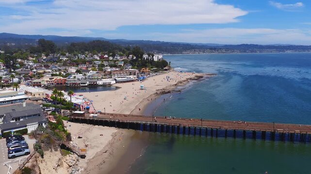 Aerial view of Capitola, California, showing a wooden pier, sandy beach with umbrellas, coastal buildings, and gentle ocean waves under a clear sky.