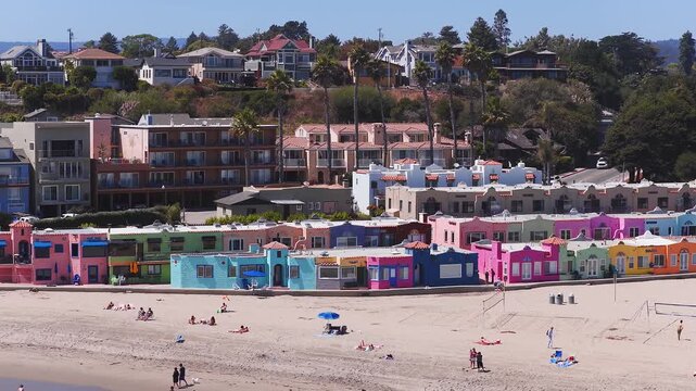 Aerial view of Capitola, California, showing colorful beachfront cottages, a sandy beach with visitors, and two bridges on a sunny day.