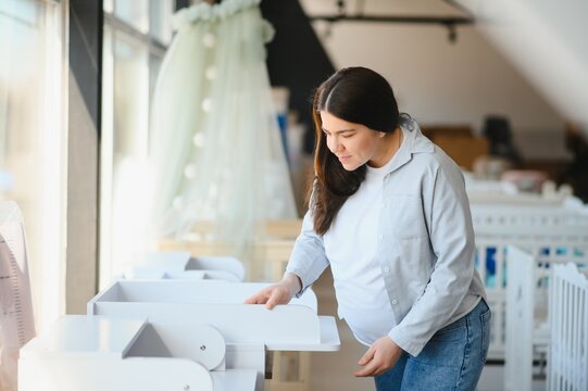 Pretty pregnant woman choosing changing table for newborn in infant shop - Powered by Adobe