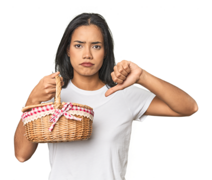 Latina holding a wicker basket showing a dislike gesture, thumbs down. Disagreement concept.