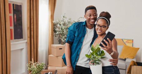 In their new apartment, a girl stands with a plant in her hands, ready to place it on the windowsill. A boy approaches her and raises his phone, smiling as he takes a selfie.