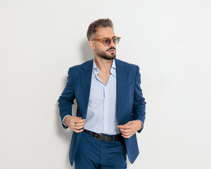 cool elegant man with sunglasses buttoning navy blue suit and looking to side