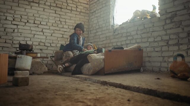 wanderer girl in worn clothes and beanie tends to sick child lying on mattress inside cold abandoned brick room, cookware scattered on floor, faint sunlight through window