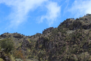 Rugged mountain landscape beneath a brilliant blue sky, featuring steep cliffs, jagged rock formations, and sparse vegetation clinging to the terrain