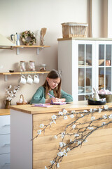 A happy girl writes in her notebook at a modern kitchen island, surrounded by cozy Easter and spring decorations, including cherry blossoms and decorative eggs. Perfect for themes of childhood, creati