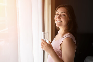 Pregnant woman drinking water and smiling by the window