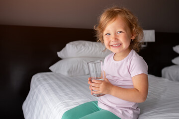 Smiling girl drinking water glass sitting on bed hotel room