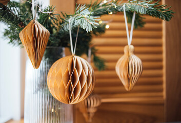 Decorative honeycomb paper ornaments in warm tones hanging from a pine branch, creating a cozy holiday atmosphere with a wooden background and soft lighting