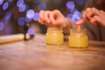 Hands of a person carefully stirring wax in glass jars on a rustic wooden table, with soft bokeh lights in the background, creating a cozy crafting atmosphere
