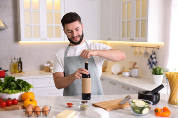 Smiling man opening bottle of wine while cooking at table in kitchen