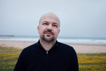 Bald man with beard stands on beach, wearing black sweater, gazing thoughtfully at ocean waves, surrounded by green grass and yellow flowers, capturing serene coastal atmosphere