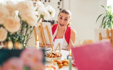 Female artist with brown hair, wearing an apron, is smiling at the camera in a bright studio filled with flowers, easels, and art supplies, showcasing a creative atmosphere
