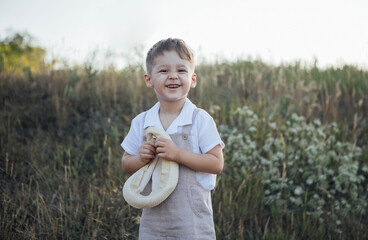 Young boy with light brown hair, wearing a white shirt and beige overalls, joyfully holding a soft toy in a grassy field, surrounded by wildflowers and natural scenery