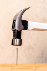 A metal hammer with a white handle drives a nail into a wooden board, illustrating the process of construction and working with tools