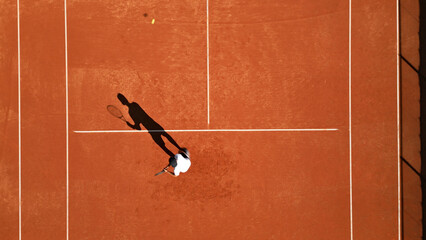 Flat lay photo of tennis player practicing on clay court, mid-action with dynamic shadow. Highlights sports training, skill development, textured clay, and athletic movement.