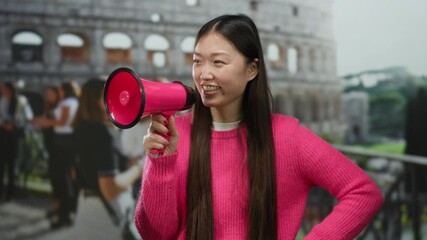 Woman smiling holding megaphone near roman coliseum wearing pink sweater vibrant city backdrop lively energy outdoor interaction speaking engagement.