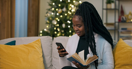 Winter evening. Beautiful african girl sitting on sofa in living room and reading book in blue...