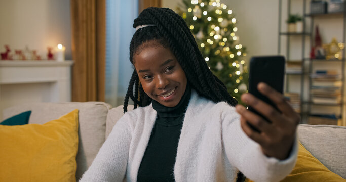A young student of multi-ethnic background sits on a sofa, taking a selfie. Dressed in a black golf and white cardigan, she takes photos and chooses the best. A Christmas tree flickers behind.