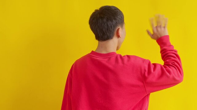 Rear view of Young man waving hello, a friendly gesture of greeting isolated on yellow background