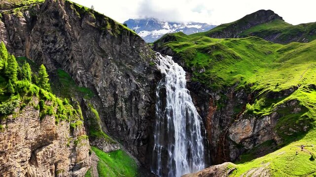 Engstligenfalle waterfall thunders down a rugged cliff amid green hills and snow capped Swiss Alps, sunlit valley scene, Adelboden, Switzerland, drone pulling out
