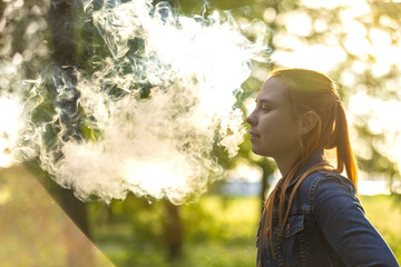 An young woman with long, red hair vaping in a forested area. The person is captured from the side, exhaling a thick cloud of vapor from an electronic cigarette. Summer landscape in a background.