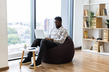 Man sitting on beanbag indoors working on laptop in office with large windows. Modern interior...
