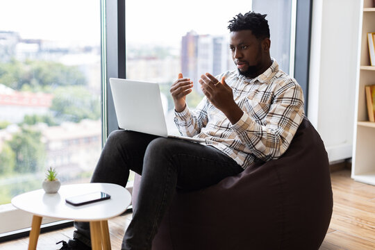 Young adult African American man sitting on bean bag using laptop for video call, communicating confidently in modern room with bookshelf and window view, showcasing work from home - Powered by Adobe