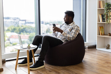 Young adult male sitting on brown beanbag, using smartphone and laptop, modern leisure concept in bright indoor setting.