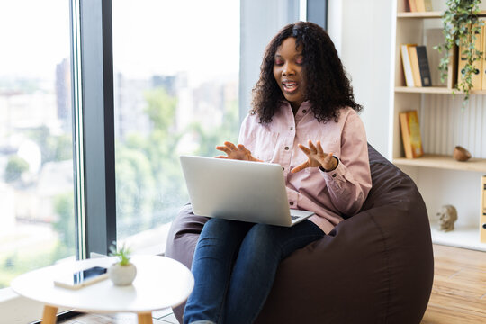 Young African American woman smiling and using laptop in casual setting during video chat. Comfortable atmosphere with natural light, bean bag seating.