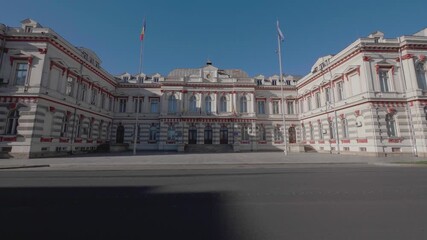 A cinematic, slow-motion dolly shot moves across a public square towards the historic Administrative Palace in Bacau, Romania, a grand neoclassical government building. - Powered by Adobe