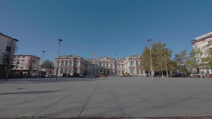 A cinematic, slow-motion dolly shot moves across a public square towards the historic Administrative Palace in Bacau, Romania, a grand neoclassical government building. - Powered by Adobe