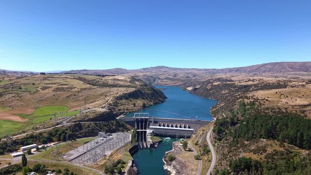 Drone flies toward Roxburgh hydroelectric dam, capturing the power station, full dam structure, and scenic Roxburgh Lake in Central Otago, New Zealand.