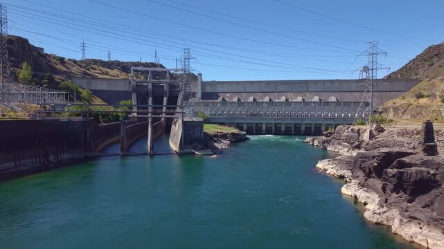 Aerial drone shot flying in reverse low over the Clutha River, revealing the full span of Roxburgh hydroelectric dam in Central Otago, New Zealand.