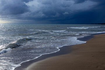 Stormy Mediterranean Sea at sunset with dramatic clouds and sun rays breaking through, waves washing sandy beach, silhouettes of swimmers in distance. Side, Antalya, Turkey, March.
