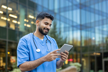 Young male doctor smiling while looking at and using a digital tablet, wearing medical scrubs and a...