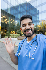 Indian male doctor in blue scrubs with stethoscope smiling and waving while taking a selfie outside...