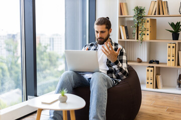 Young adult male seated on bean bag video chatting using laptop, bright modern home interior with natural window light