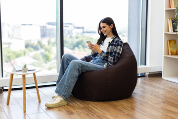 Young adult woman seated on bean bag in modern lounge environment using smartphone. Scene shows casual lifestyle and leisure time indoors. Well-lit surroundings with natural light and wooden flooring.