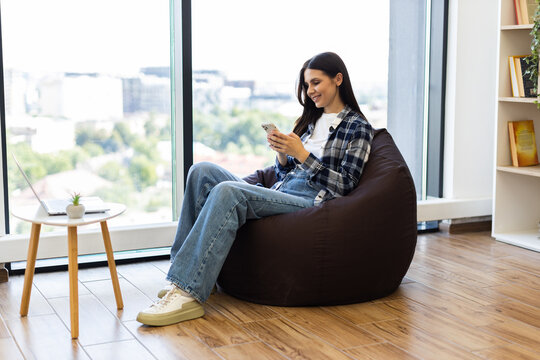 Young adult woman seated on bean bag in modern lounge environment using smartphone. Scene shows casual lifestyle and leisure time indoors. Well-lit surroundings with natural light and wooden flooring.