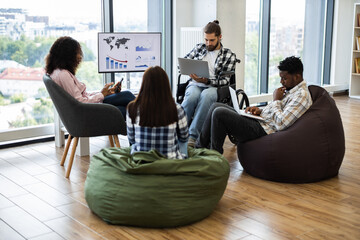 Group of young adults, diverse gender and race, working together in modern casual office. Collaborative atmosphere, bean bags, wheelchair accessibility, and digital devices convey teamwork, inclusion.