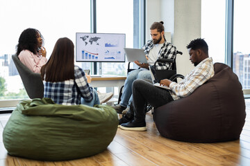 Group of young adults, diverse gender and race, working together in modern casual office. Collaborative atmosphere, bean bags, wheelchair accessibility, and digital devices convey teamwork, inclusion.