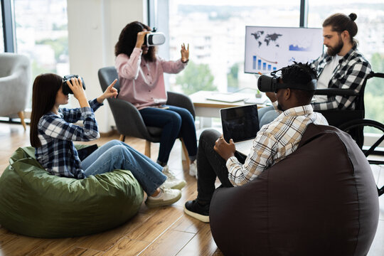 Group of diverse adults using virtual reality headsets and laptops in modern office setting. Individuals of various backgrounds collaborating in technology-driven workspace fostering creativity