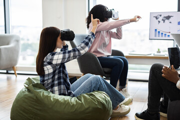 Two young women using VR headsets in modern office. One sits on bean bag others engage with technology. Mixed environment promoting innovative collaboration.