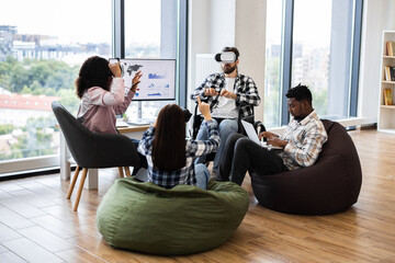 Young adults engage with virtual reality devices and laptops in collaborative office. Diverse teams demonstrating technology interaction seated on floor and chairs creating innovative atmosphere.