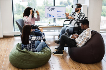Diverse group using VR glasses and laptops in teamwork collaboration in office environment. Interacting with technology while seated on bean bags and chairs, showcasing inclusivity