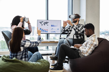 Diverse group using VR glasses and laptops in teamwork collaboration in office environment. Interacting with technology while seated on bean bags and chairs, showcasing inclusivity