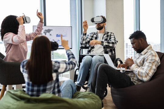 Diverse group of young adults including male wheelchair user enjoying virtual reality. Another male working on laptop sitting on bag chair in modern office setting
