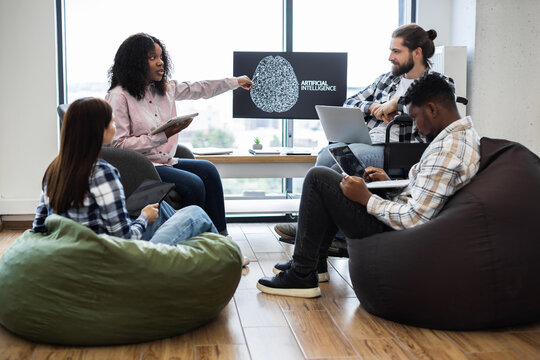 Group of men and women collaborating on artificial intelligence presentation in relaxed office. Includes professionals with diverse backgrounds, surrounded by beanbags, using digital technology