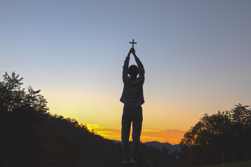 Woman holds a cross and raises her hands to the sky praying to God at sunset background