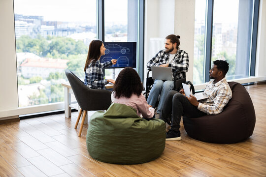 Diverse group of young engineers collaborating on car design project using digital tools. Office features large windows, comfortable seating. Participants are varied in gender, race, ages